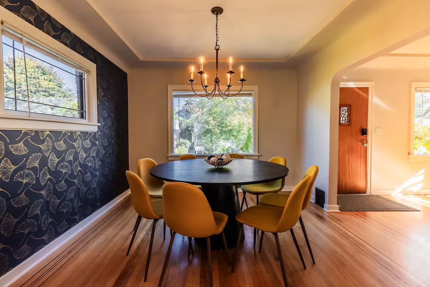 A dining area with a round table, yellow chairs, a chandelier, and a window with a view of trees.