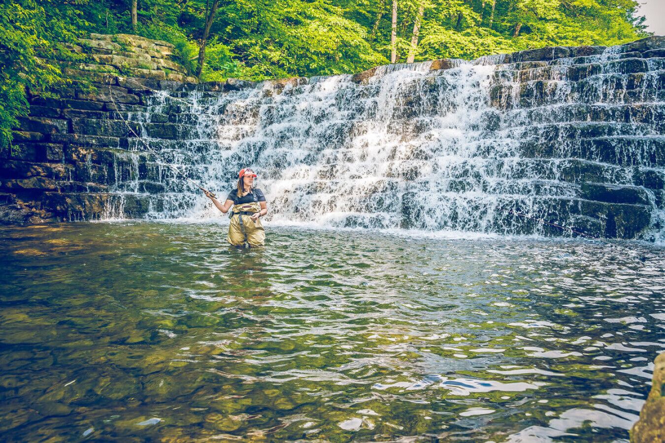A person standing in a river with a waterfall in the background.