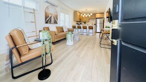 A modern living room with a sofa, coffee table, and decorative plants. A kitchen area with a refrigerator and dining table is visible in the background. - 4173-Newly renovated stylish home (San Diego)