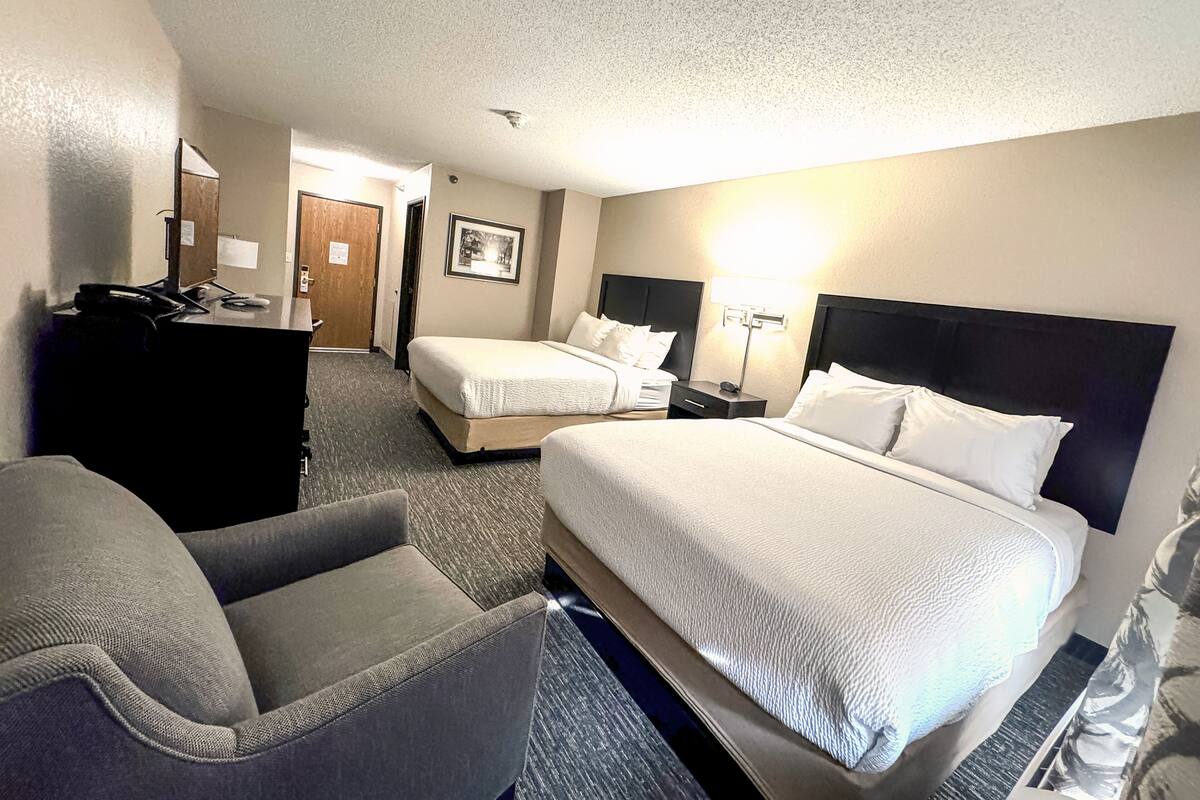 A hotel room with two beds, a desk, a chair, and a lamp.