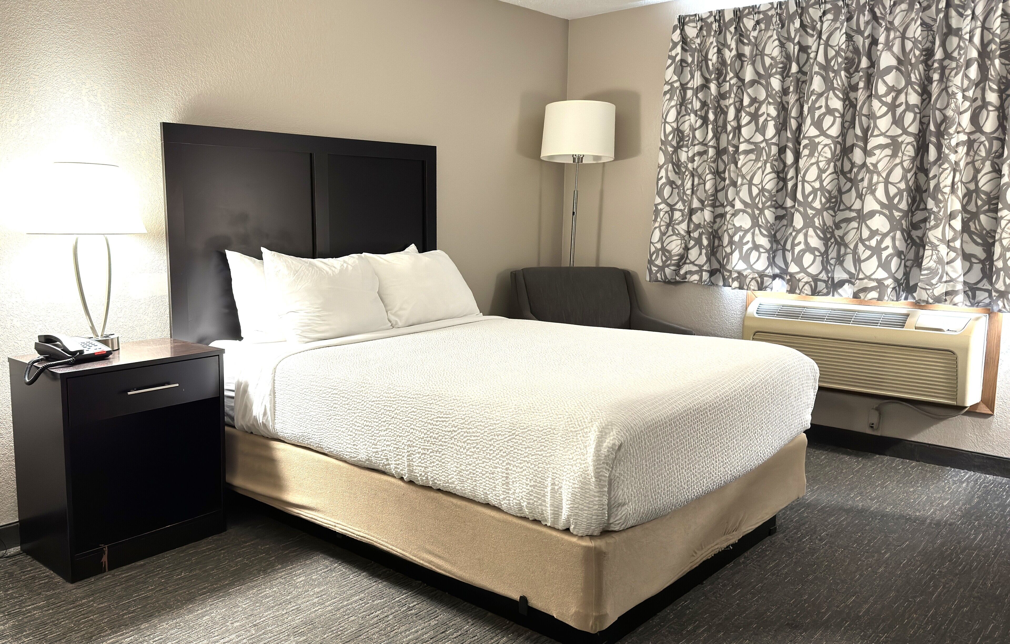 A hotel room with a bed, a nightstand with a lamp, a chair, a wall-mounted air conditioner, and a patterned curtain.
