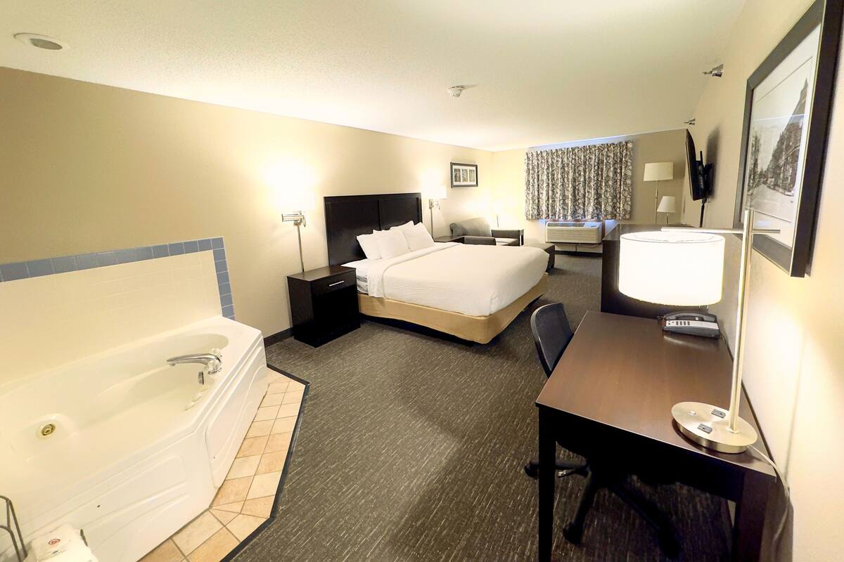 A hotel room with a large bed, a desk, a chair, a desk lamp, a TV, a window with curtains, a wall-mounted TV, a wall-mounted picture, a bedside table, a lamp, and a bathtub.