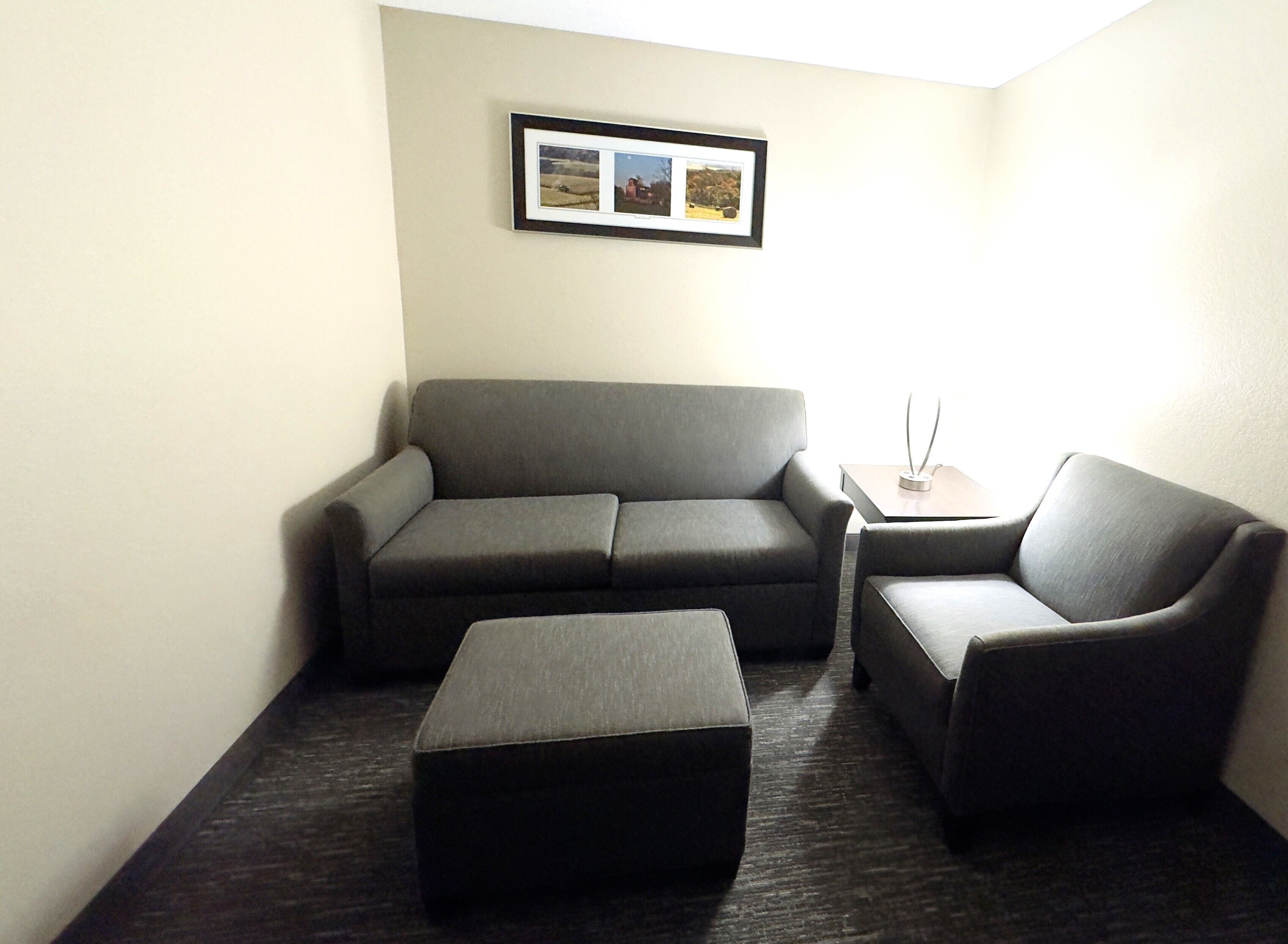 A modern living room with a grey sofa, a matching armchair, and a small ottoman. There is a framed picture on the wall and a small table with a vase.