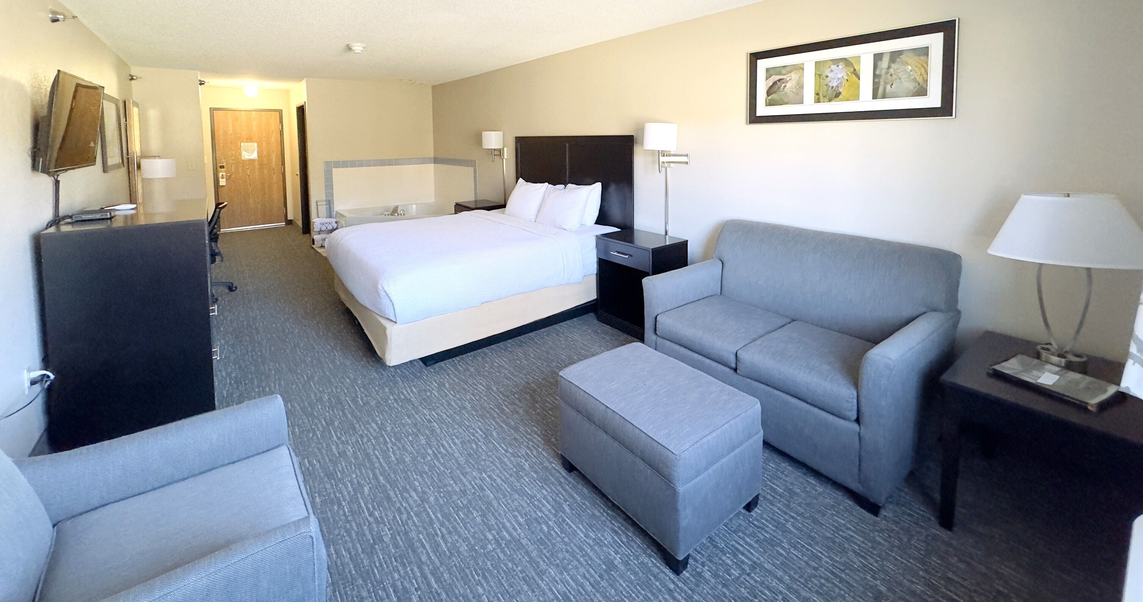 Een hotelkamer met een bed, een grijze fauteuil, een klein poufje, een bureau met een lamp en een lijst met een afbeelding aan de muur.