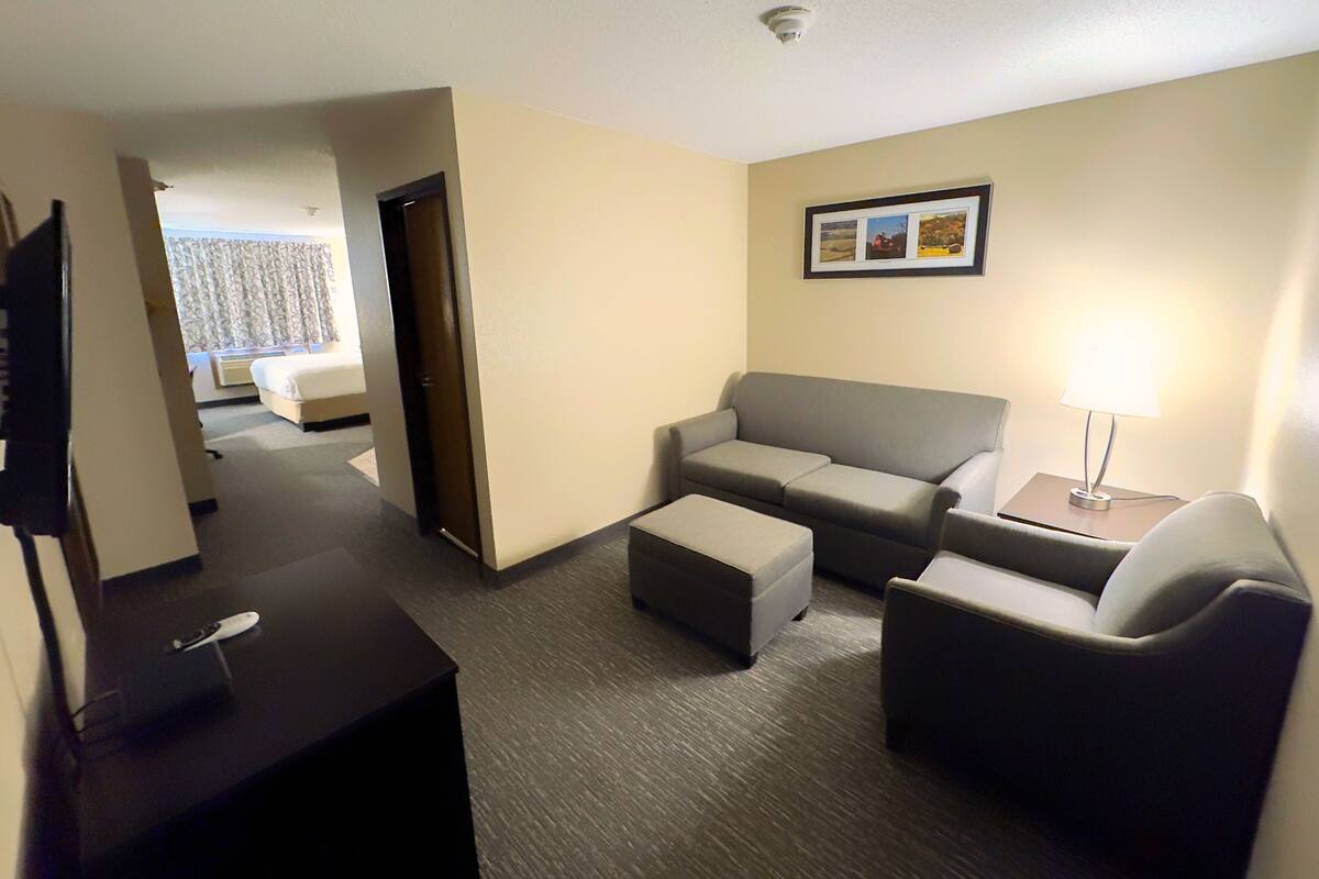 A hotel room with a sofa, two armchairs, a small table with a lamp, a TV on a stand, and a bed in the background.