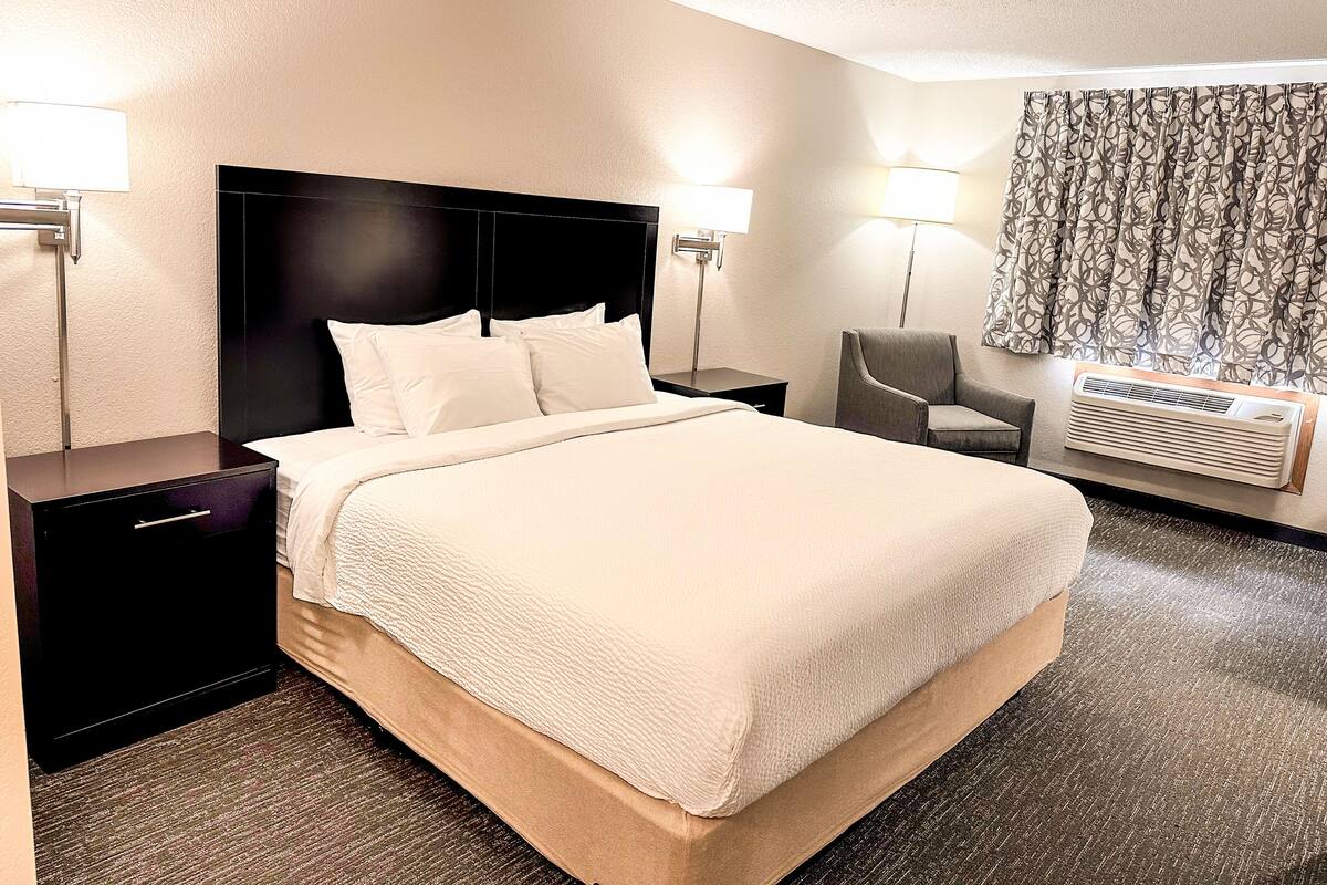A hotel room with a large bed, two bedside tables, a chair, and an air conditioning unit.