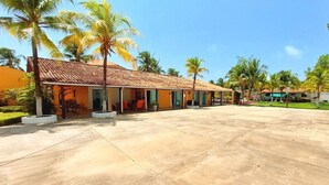 A row of single-story buildings with a tiled roof, surrounded by palm trees and a paved area. - Aconchego do Caranã (Salinópolis)