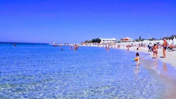Una playa con agua cristalina, gente y edificios al fondo.