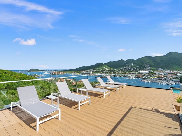 A deck with white lounge chairs overlooking a marina with boats and a mountainous backdrop.