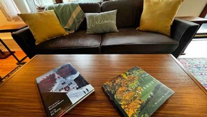 Una mesa de centro de madera con dos libros, un sofá de cuero oscuro con cojines, y una ventana con cortinas al fondo.