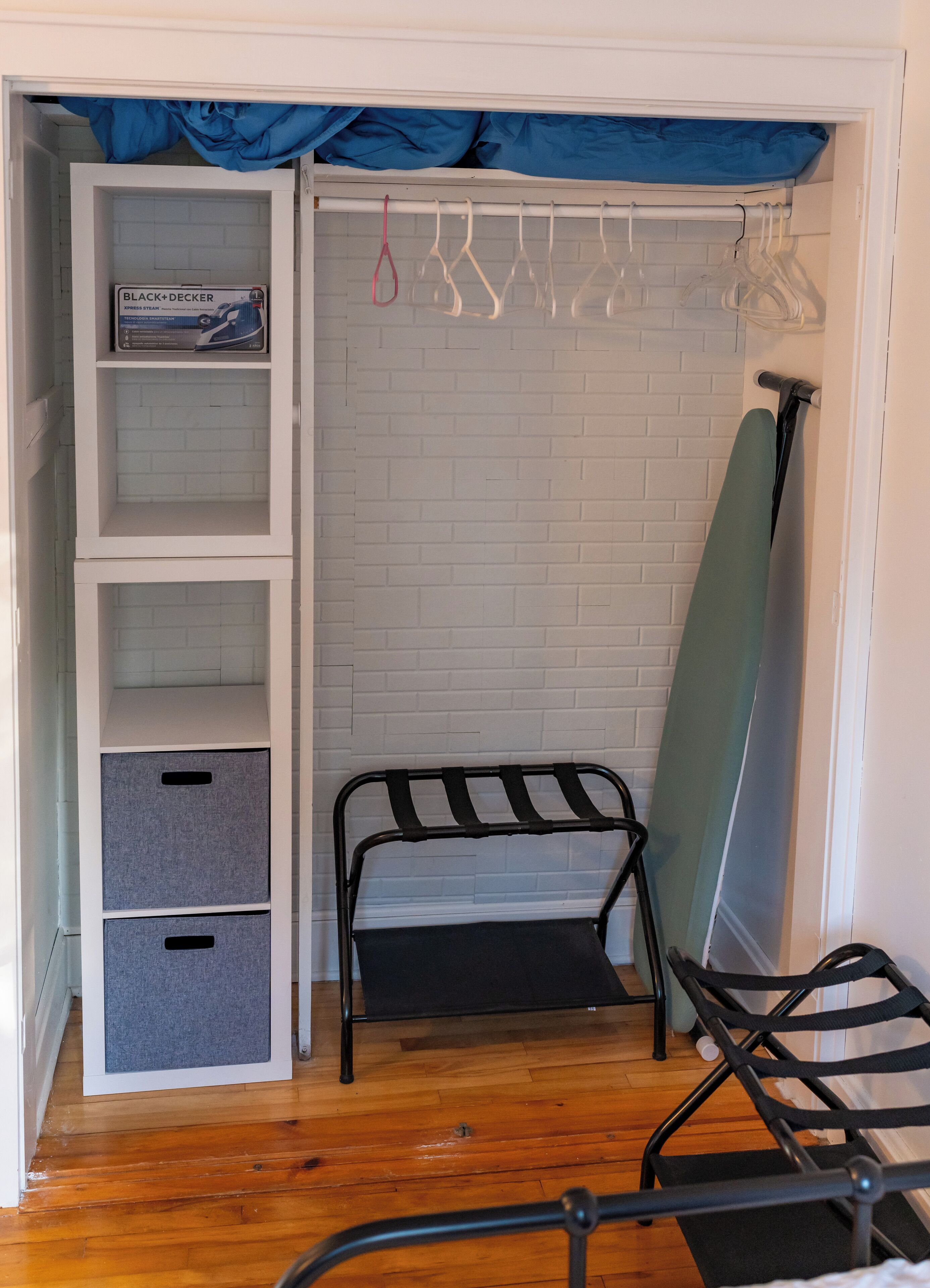 A room with a white brick wall, a wooden floor, a metal bench, a storage unit with grey bins, a blue curtain, and a hanging rack with clothes.