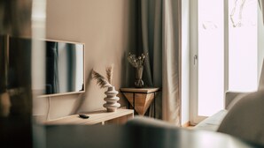 A modern room with a flat-screen TV, a wooden side table, a vase with dried flowers, and a view of the outdoors through a large window. - Luga Homes - Karli (Leipzig)