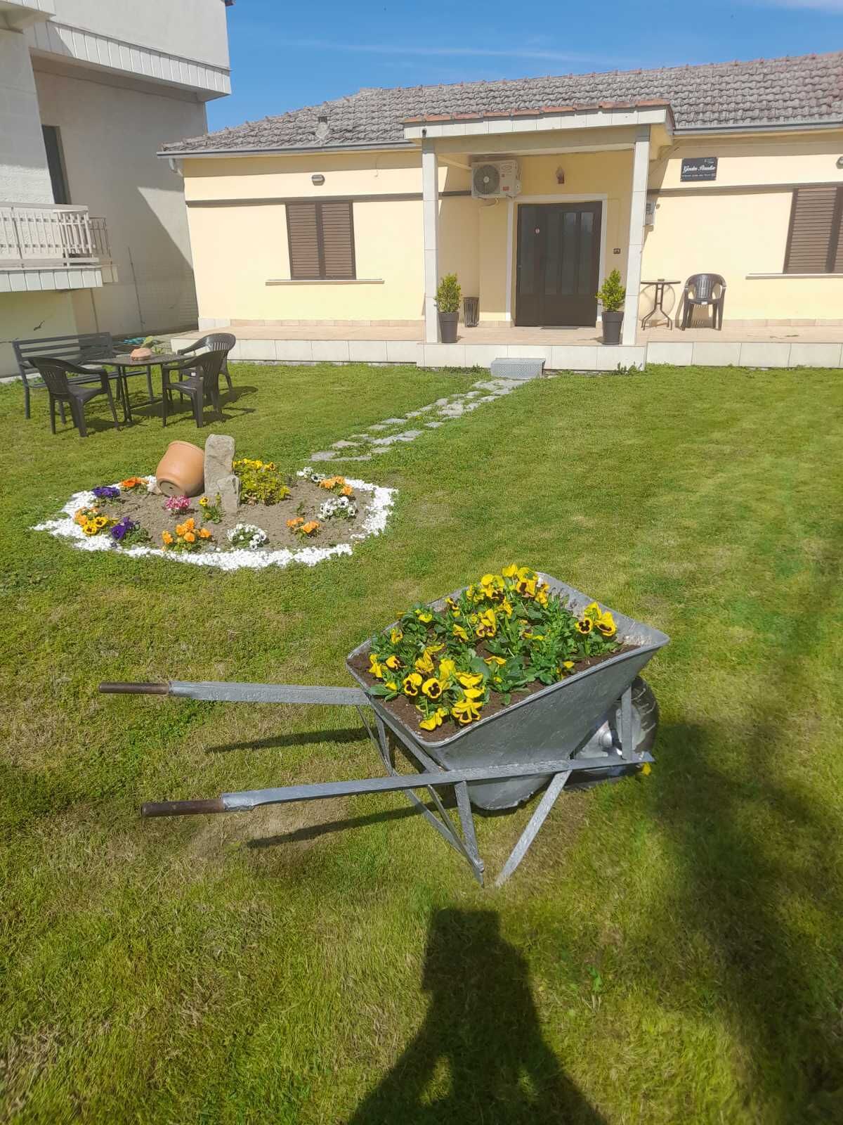 Una casa con un camino empedrado que lleva a la entrada, un carretón con flores amarillas y mobiliario de exterior.