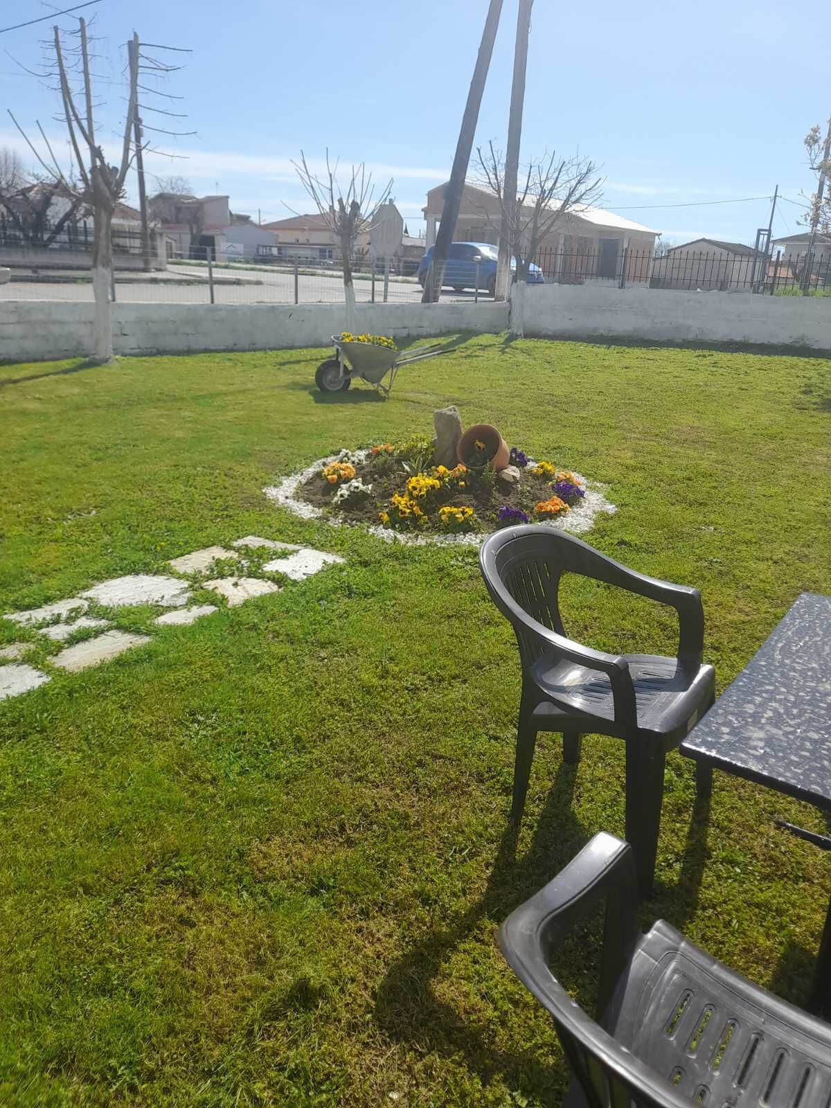 Un espacio exterior con césped, un pequeño macizo de flores, un carretilla y una mesa con sillas.