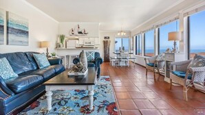 A spacious living area with a black leather sofa, a coffee table, and a dining area with wicker chairs. A kitchen is visible in the background, and large windows offer a view of the ocean. - Quick walk to the Beach
3 Bedrooms 1.5 Bath
 Sleeps 6 (Dillon Beach)