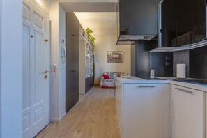 Un pasillo estrecho con una zona de cocina que incluye armarios blancos, una estufa empotrada y una pequeña sala de estar con un cojín rojo.
