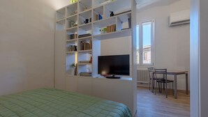 Quarto com uma cama, televisão, escrivaninha com cadeiras e estante de livros.