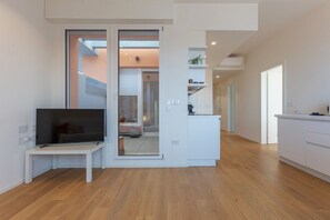 A modern living space with a TV on a stand, a sliding glass door leading to a balcony, and a kitchen area with white cabinets.