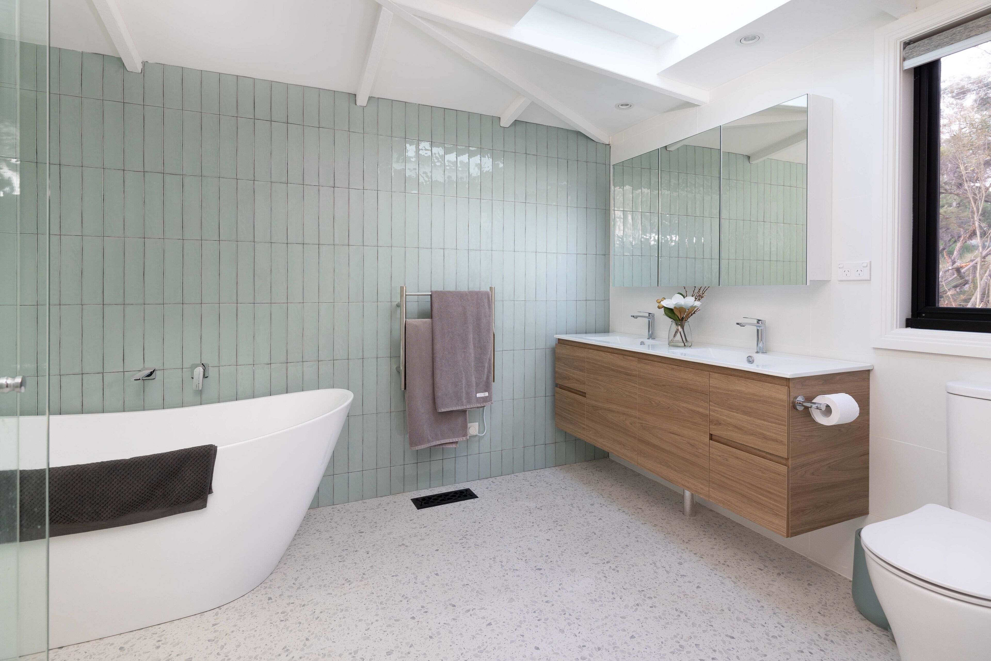 A modern bathroom with a freestanding bathtub, a wooden vanity, a large mirror, and a window.