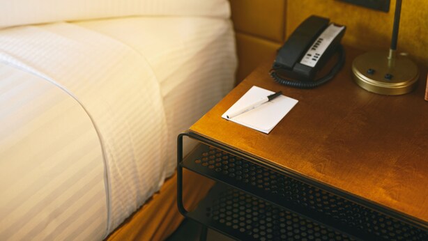 A neatly made bed with white linens, a bedside table with a lamp, a phone, and a notepad. - Hotel Julian (San Francisco)