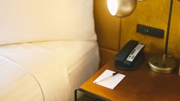 Una cama bien hecha con sábanas blancas, una mesita de noche con una lámpara, un teléfono y un bloc de notas.