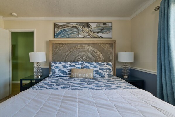 A hotel room with a bed, two bedside tables with lamps, a headboard with a decorative panel, a painting above the bed, and a green door. - MillionDollarView-Oceanfront 1BR-Updated! (Salter Path)