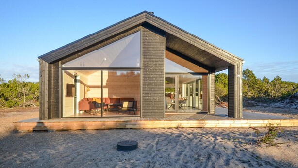 A modern, single-story wooden cabin with a large glass front, a wooden deck, and a sandy surrounding. - 3 bedroom lovely home in Anholt (Anholt)