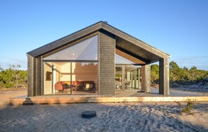 Eine moderne, eingeschossige Holzhütte mit grosser Glassfront, einer Holzterrasse und einer sandigen Umgebung.