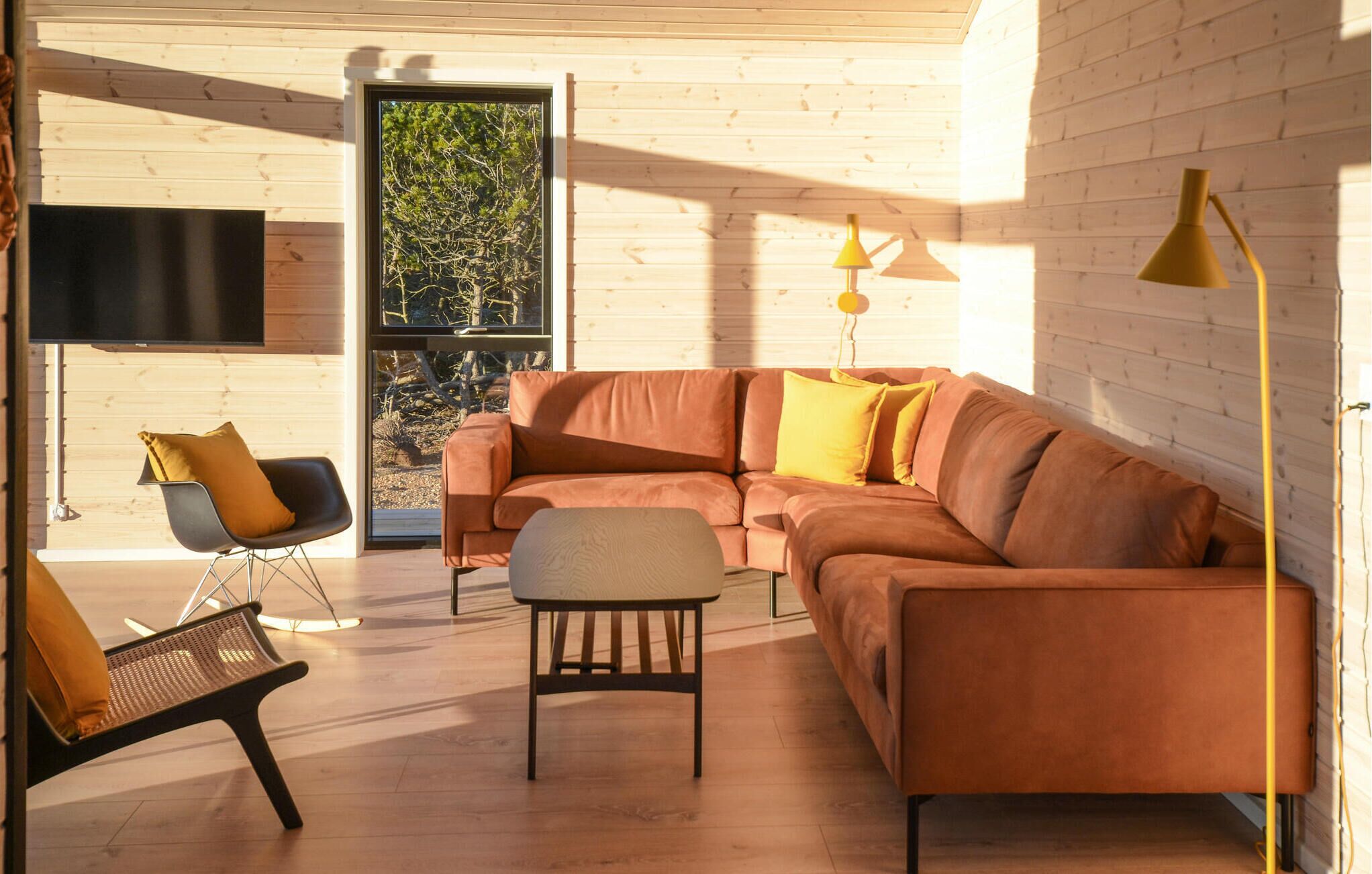 Een moderne woonkamer met een hoekbank, een bijzettafeltje en een fauteuil. Er is een televisie aan de muur bevestigd en een raam met uitzicht op bomen.