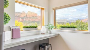 A room with two windows overlooking a mountainous landscape, a white desk with a potted plant, and a small stool.