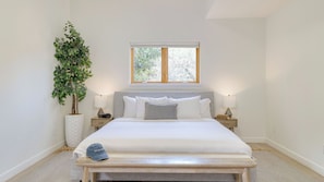 Une chambre à coucher avec un lit, des tables de chevet, une fenêtre, une plante et un banc.