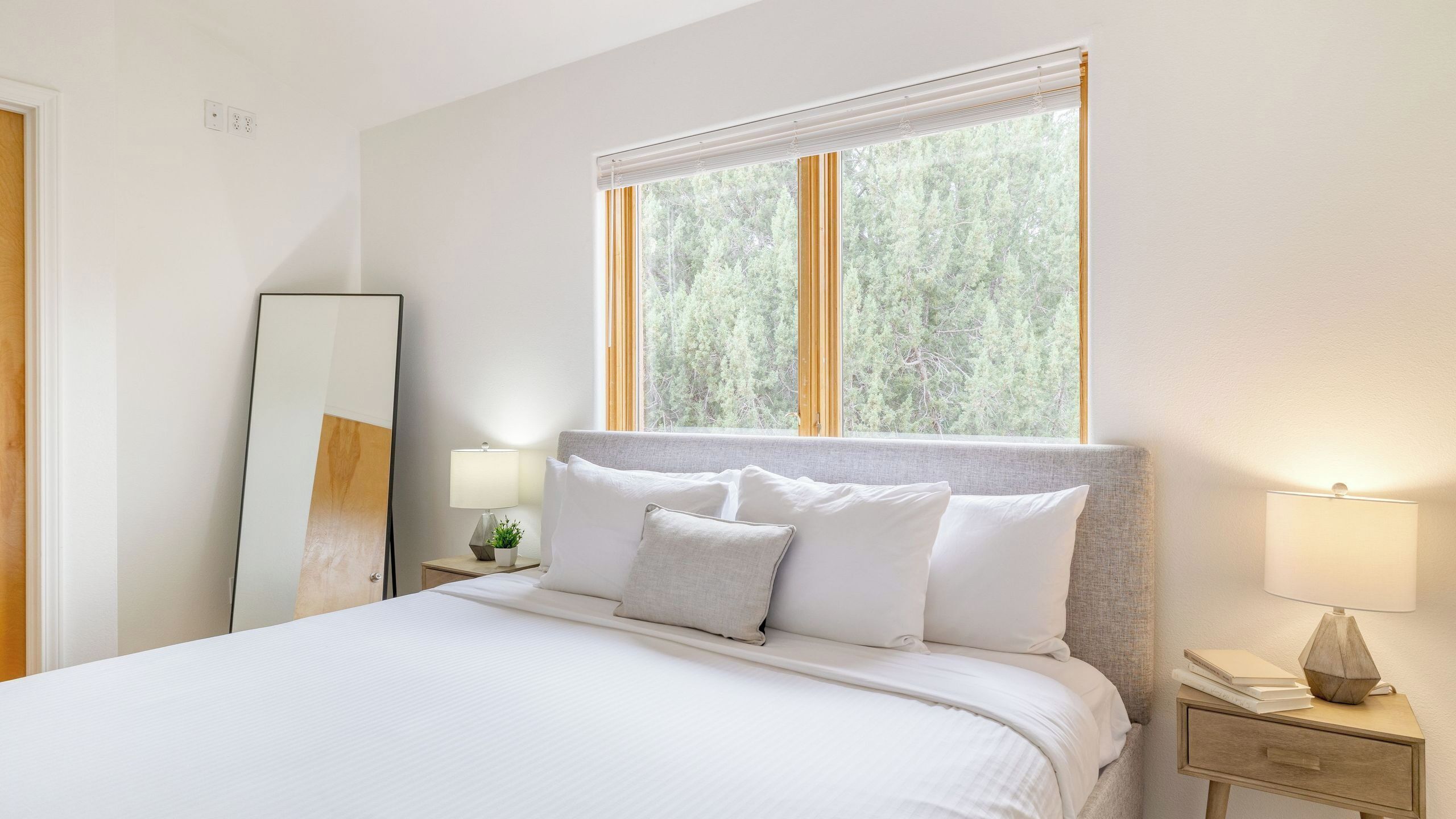 A neatly made bed with white linens, a bedside lamp, a small table with a plant, and a window with a view of trees.