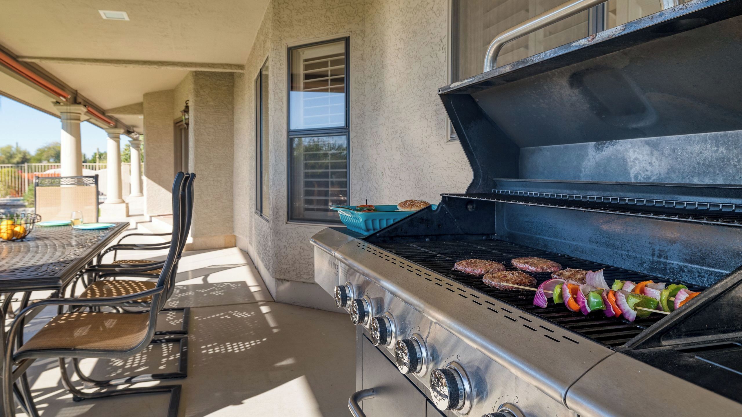 Un barbecue extérieur avec des hamburgers et des légumes, une table avec des chaises et une fenêtre.