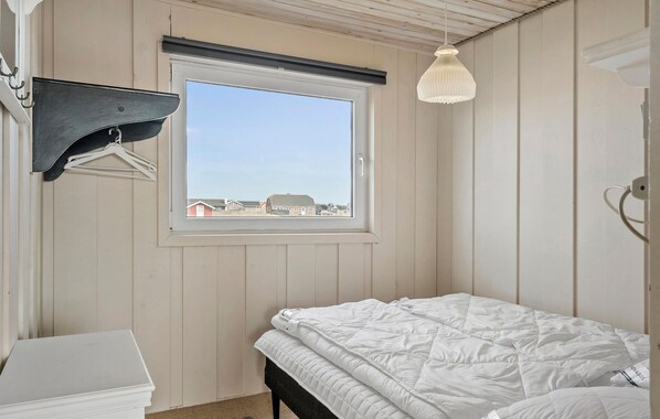 A bedroom with a bed, a window with a view of buildings, a hanging light fixture, and a coat rack with clothes.