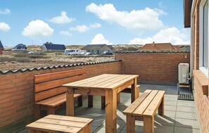 Une terrasse avec des bancs en bois et une table, un mur en briques et, en arrière-plan, une vue sur des maisons et des véhicules.