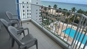 Un balcon avec des chaises en plastique, une piscine et une vue sur une plage et l’océan.