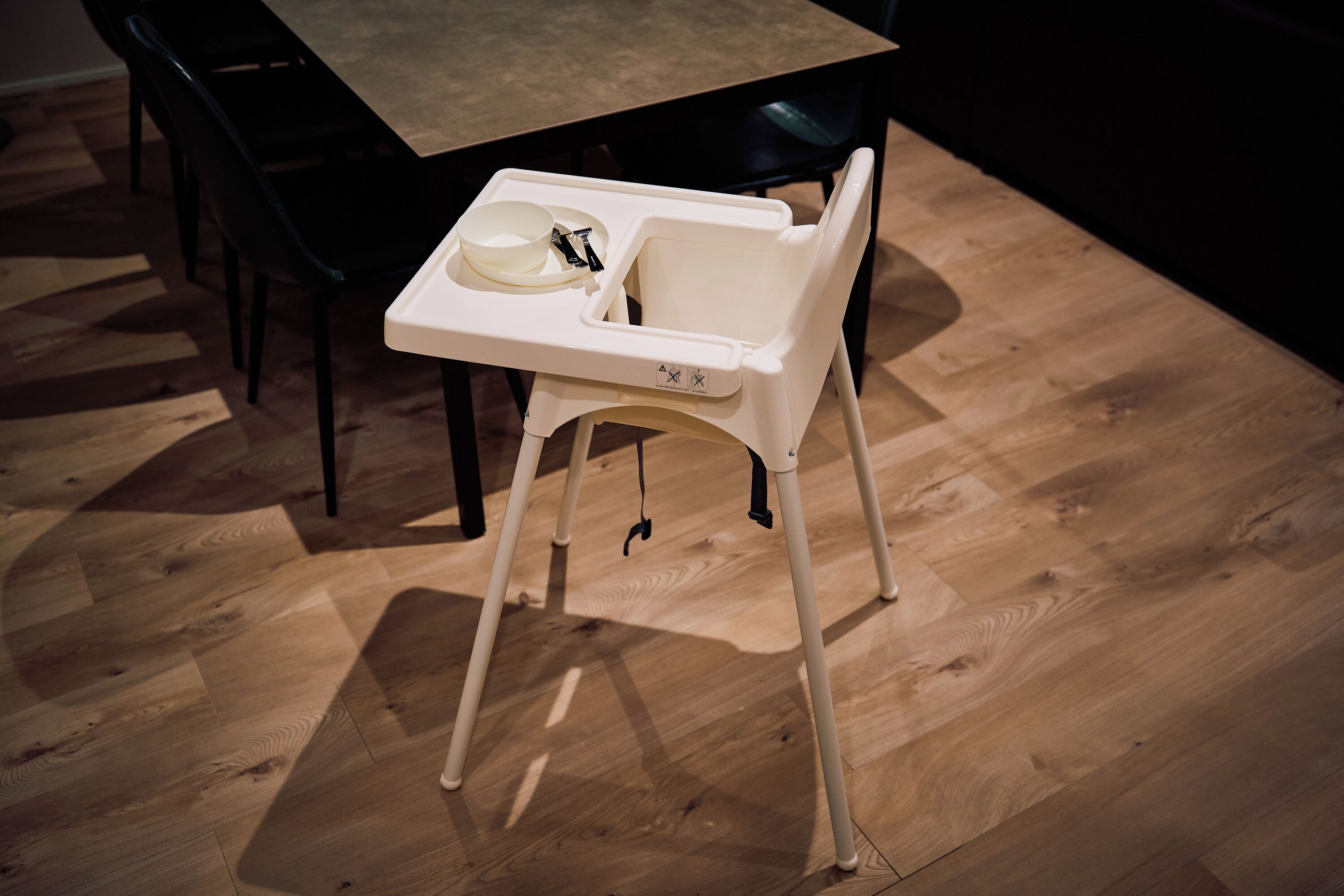Une chaise haute avec un plateau, placée sur un plancher en bois, avec une table à manger et des chaises en arrière-plan.