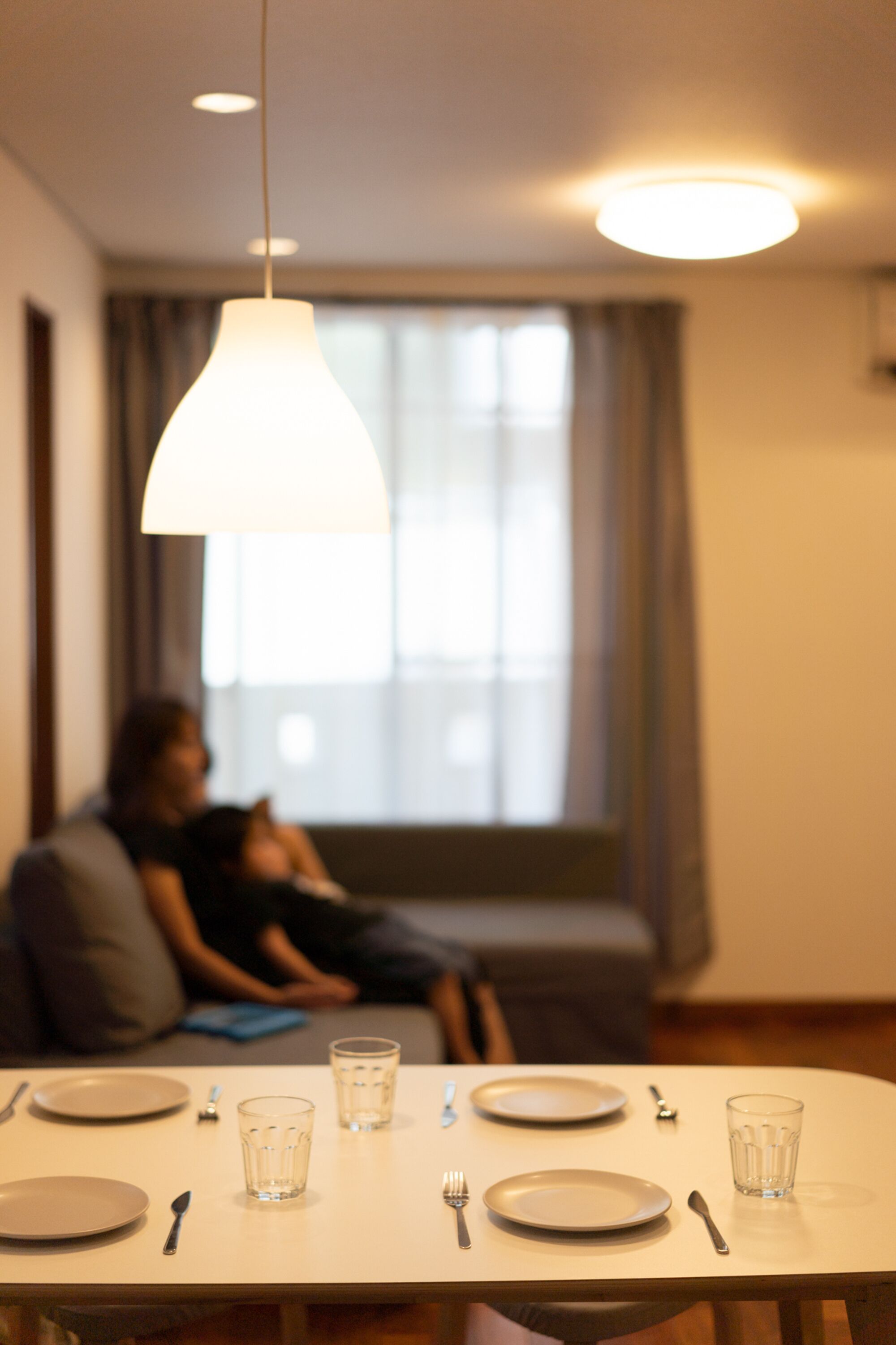 Une table à manger dressée avec des assiettes, des verres et des couverts, dans une pièce éclairée par un luminaire suspendu et dont une fenêtre est visible au fond.