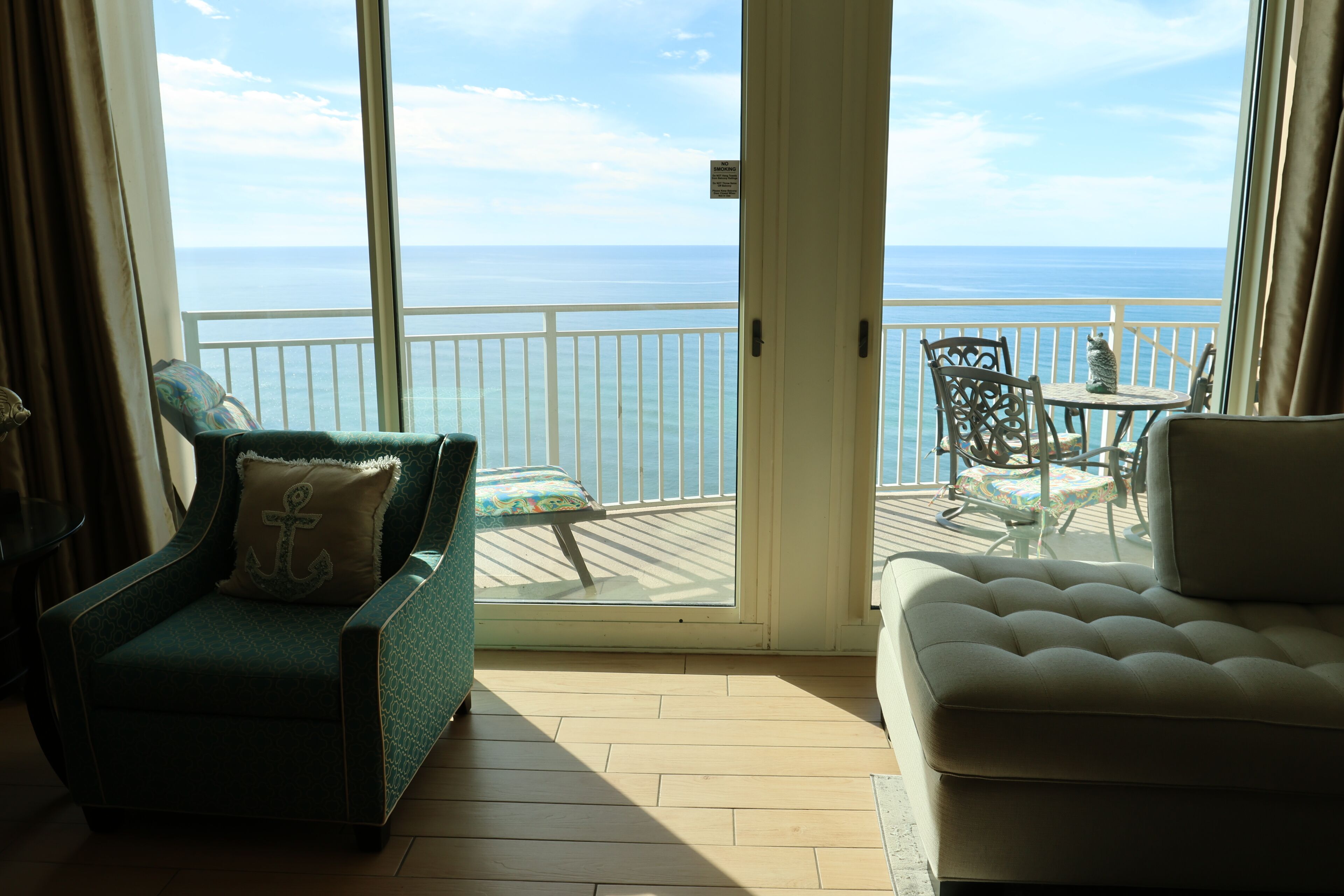 Un balcon avec vue sur l’océan, un fauteuil vert orné d’un coussin à motif d’ancre, un canapé blanc et une petite table avec des chaises.