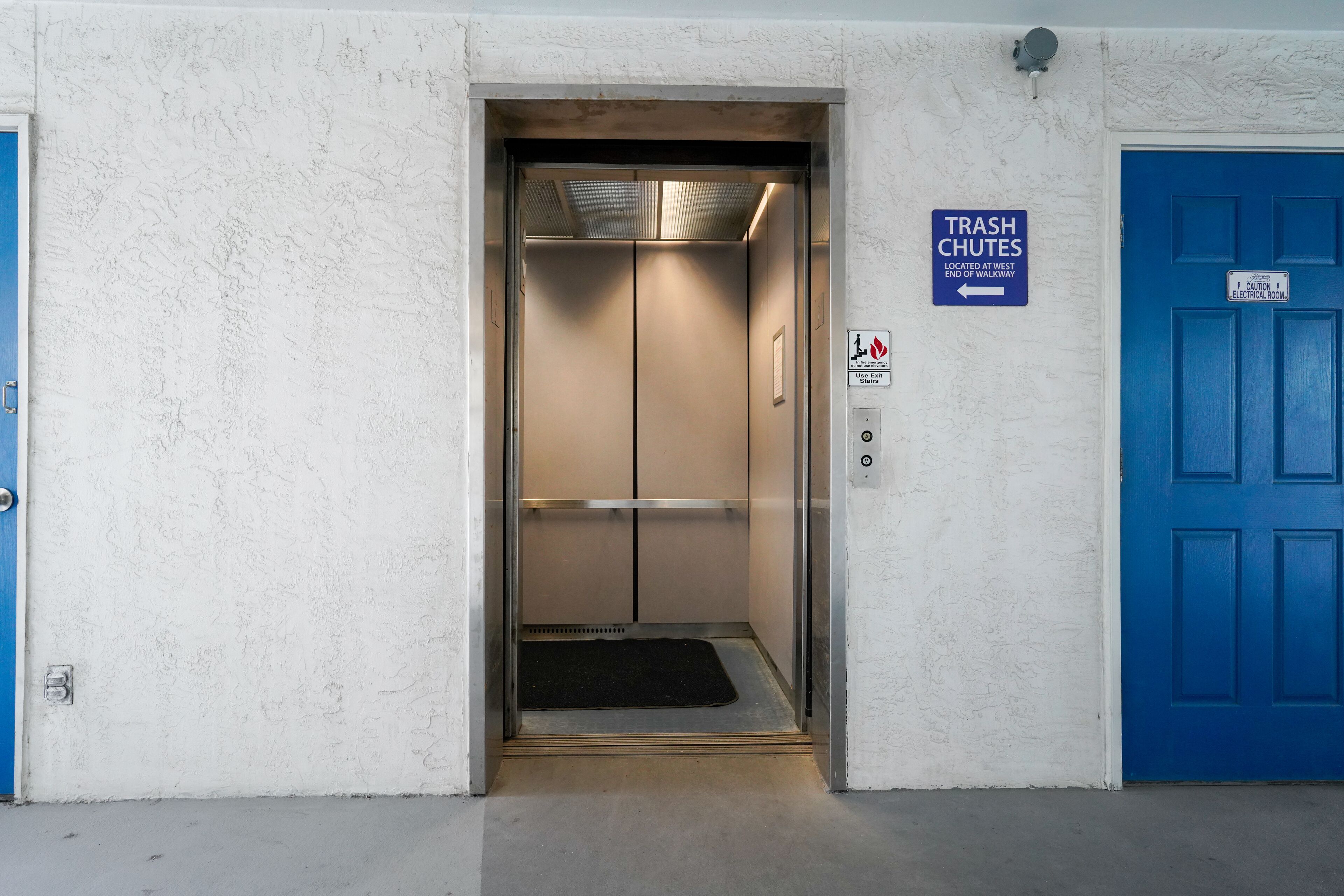 Un ascensor con un letrero de conducto de basura, una puerta azul y una señal de precaución para cuarto de electricidad.