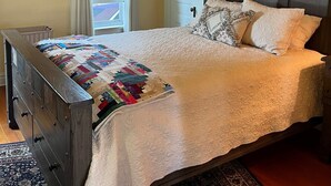 Un dormitorio con una cama de madera, colcha, cabecera oscura, puerta blanca y ventana con cortinas.