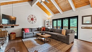 A modern living room with a sectional sofa, a flat-screen TV, a wooden ceiling, and large windows.
