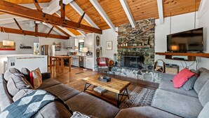 A spacious living area with a stone fireplace, wooden ceiling beams, and a modern kitchen in the background.