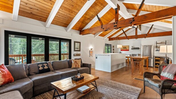 A spacious living area with a wooden ceiling, a sofa set, a coffee table, and a kitchen in the background.
