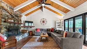 A cozy living room with a stone fireplace, a large gray sectional sofa, a wooden coffee table, and a view of the outdoors through large windows.
