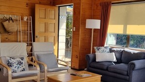 A spacious living room with wooden walls, a large window, a sofa, two armchairs, a coffee table, and a lamp. - The Bach in old Wharewaka well located for a fabulous lake holiday. (Taupo)