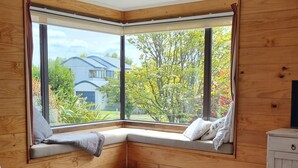 A corner room with a large window, a wooden bench with cushions, a small table with a remote control, and a white cabinet. - The Bach in old Wharewaka well located for a fabulous lake holiday. (Taupo)