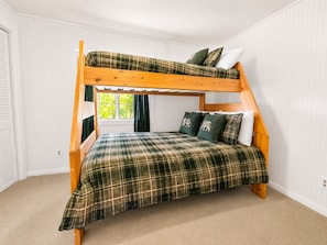 Una cama litera con colcha a cuadros y almohadas a juego, una ventana con cortinas y una pared blanca.