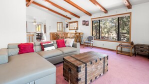A spacious living room with a large window, wooden beams, a sofa, a coffee table, and a dining area. - Silver Rays - Walk to Lake/Village, Game Room, Pool Table, Air Hockey, BBQ (Big Bear Lake)