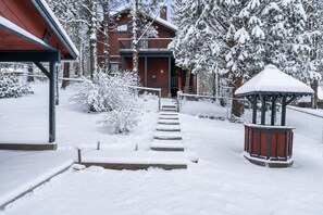 Un espacio exterior cubierto de nieve con un pabellón de madera, una escalera que lleva a un edificio y un jardín cercado.